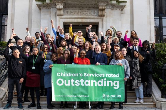 A large group of people celebrate on the steps outside Islington Town Hall holding a large green banner saying "Children's Services rated Outstanding".